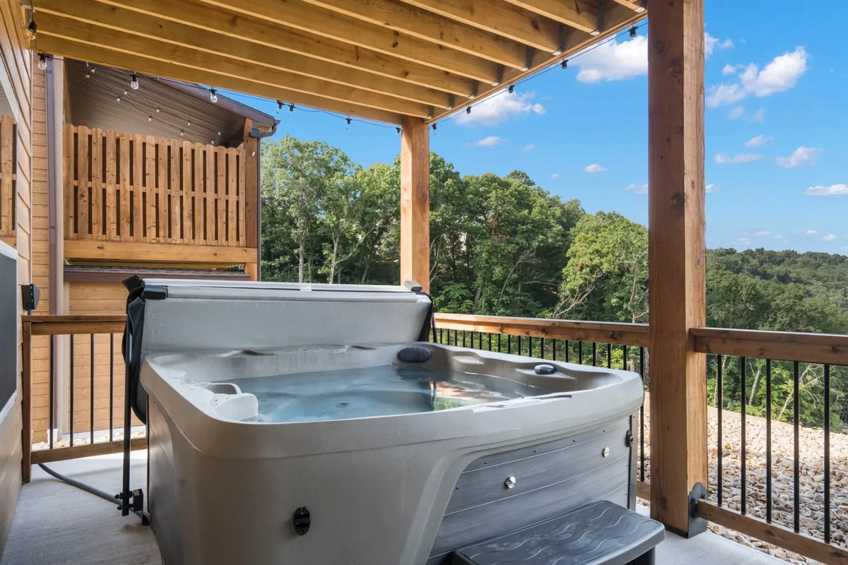 A wooden deck nestled among trees features a hot tub, inviting relaxation. The clear blue sky and the privacy provided by the surrounding natural elements make this a perfect spot to unwind after a day of adventure, emphasizing the unique 'patio with hot tub' amenity.
