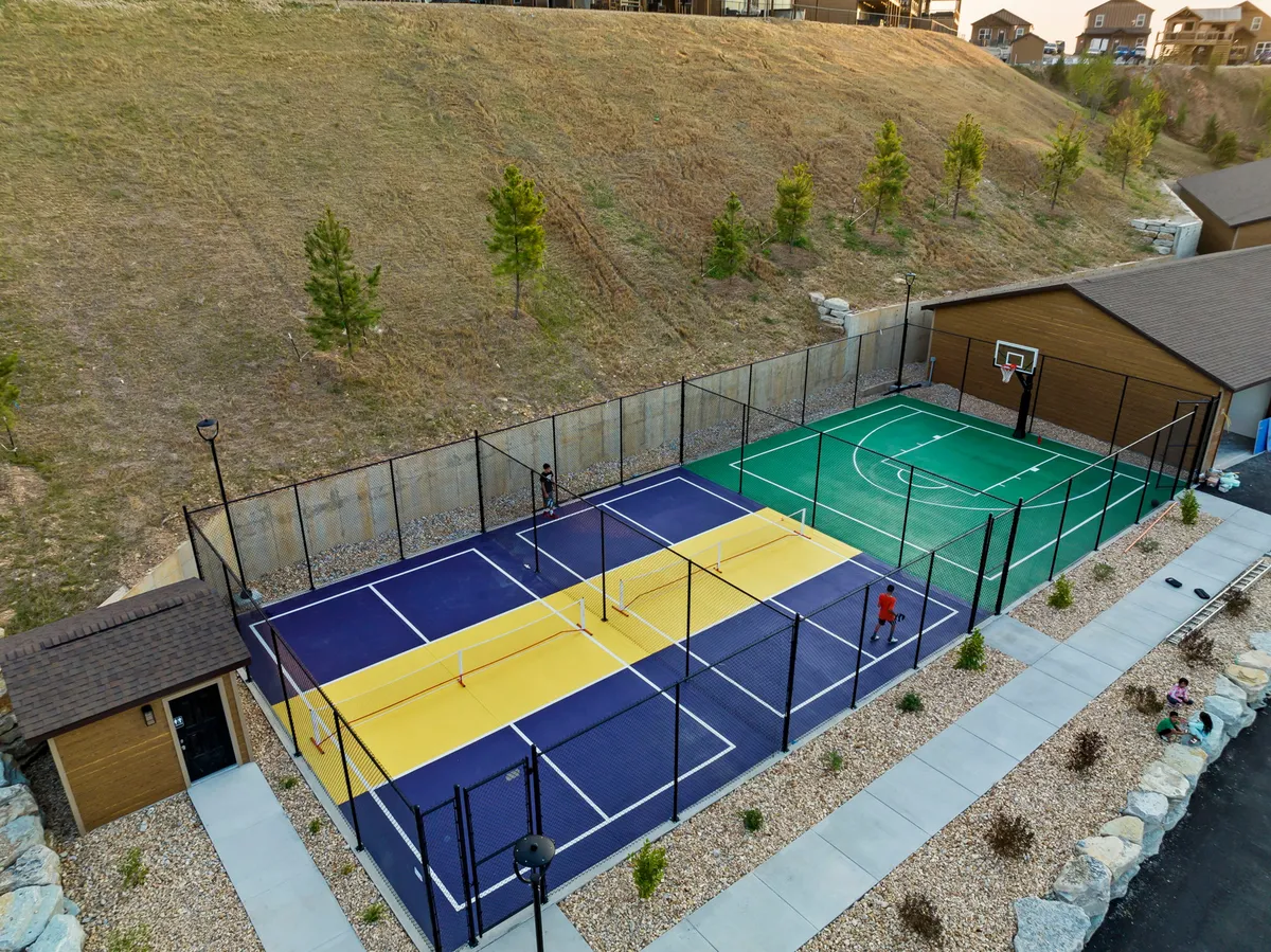 An energetic scene featuring tennis and basketball courts set amidst lush landscaping. People of all ages are seen playing and socializing, highlighting active recreation and community spirit. The courts are surrounded by open, green spaces, inviting group activities and friendly competition.