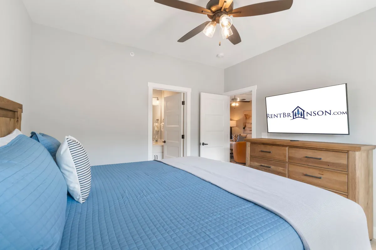 A serene bedroom featuring a bed with blue bedding, a wooden dresser, a wall-mounted TV, and open doors leading to a private ensuite bathroom. The space feels airy and comfortable, inviting relaxation and privacy.