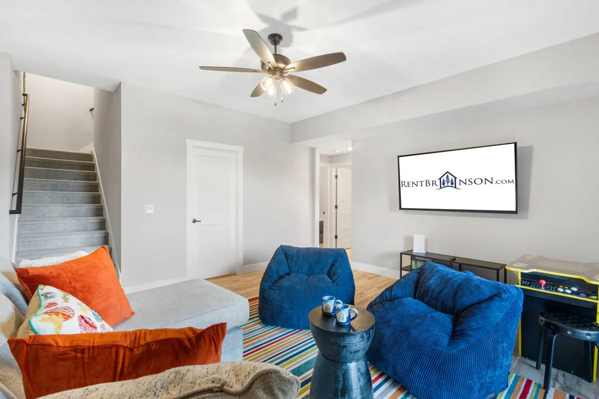 The image captures a cozy lower-level lounge space with colorful bean bag chairs, a striped area rug, a modern TV unit, and a staircase leading upward. The room exudes an inviting, relaxed vibe ideal for movie nights, socializing, and family game sessions.