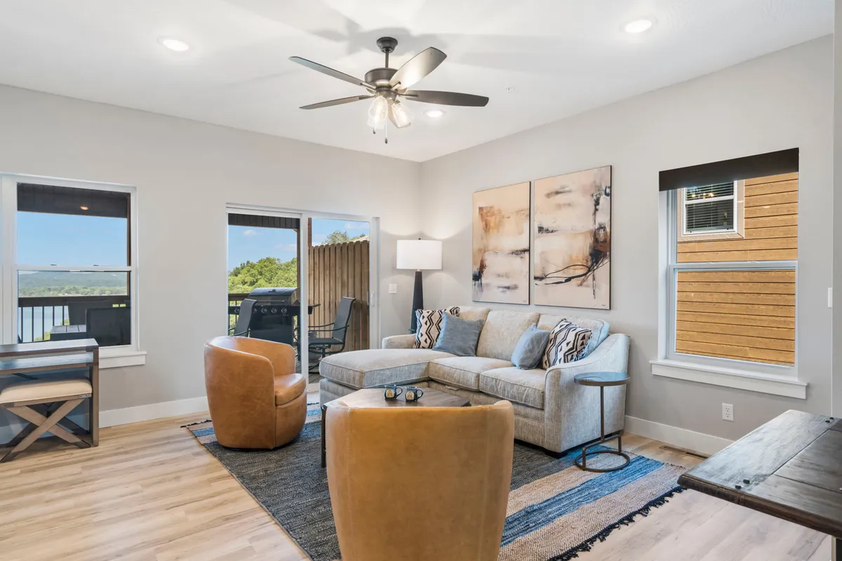 A cozy living and dining space with a stylish sofa, armchairs, wall art, and a generous dining table—all arranged near expansive windows that let in natural light and offer beautiful outdoor views. The setting invites both relaxation and shared meals.