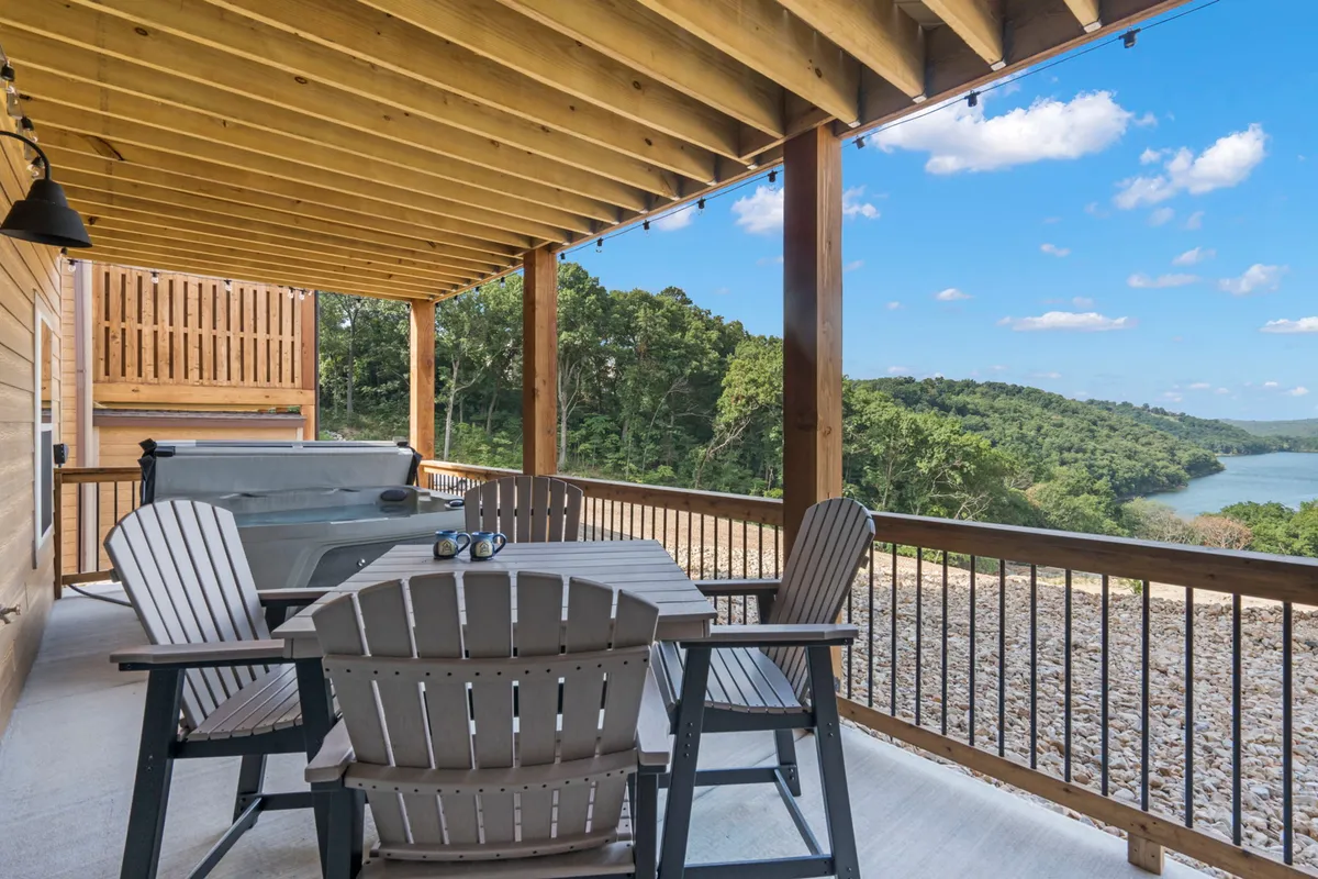A wooden deck with an outdoor dining table overlooking a river, with lush greenery in the background. The scene suggests opportunities for al fresco dining, socializing, and enjoying scenic vistas, especially as the sun sets over the water.
