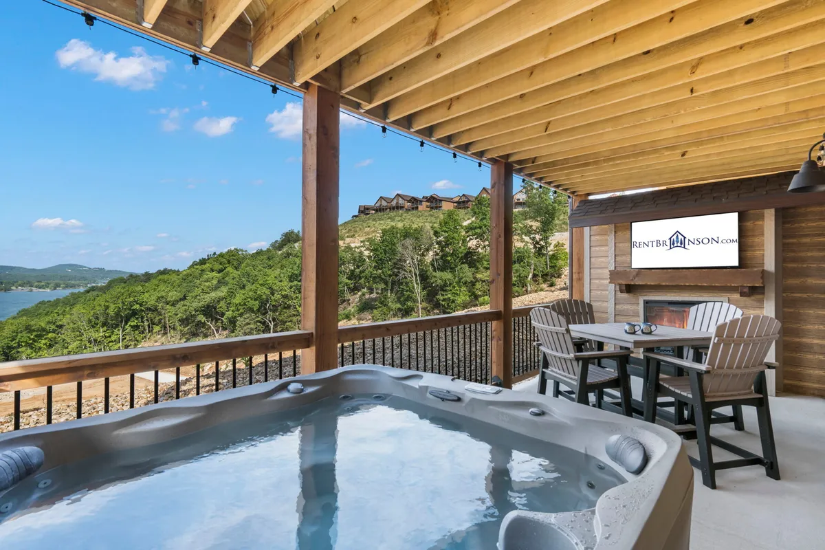 This serene exterior shot highlights a private hot tub and an outdoor dining table on a patio with sweeping lake views. The setting is tranquil, with hints of sunset light, inviting guests to soak and unwind while gazing at the Ozark landscape.