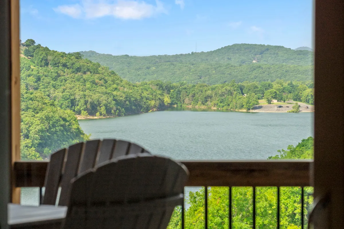 A peaceful lakeside scene featuring calm waters, forested mountains, and comfortable deck chairs arranged to enjoy the expansive view. The setting is serene, with clear skies and lush nature surrounding the lake, inviting relaxation and connection with the outdoors.